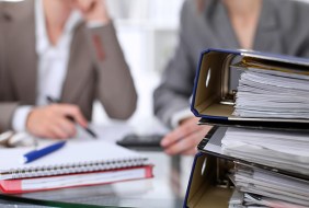 Binders with papers are waiting to be processed with businessman and secretary back in blur. Internal Revenue Service inspector checking financial document.